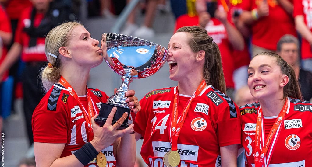 Team Esbjerg genvandt mesterskabet i 2022/2023 og her ses tre glade spillere, hvor Kathrine Heindahl deler et kys med pokalen i Blue Water Dokken
